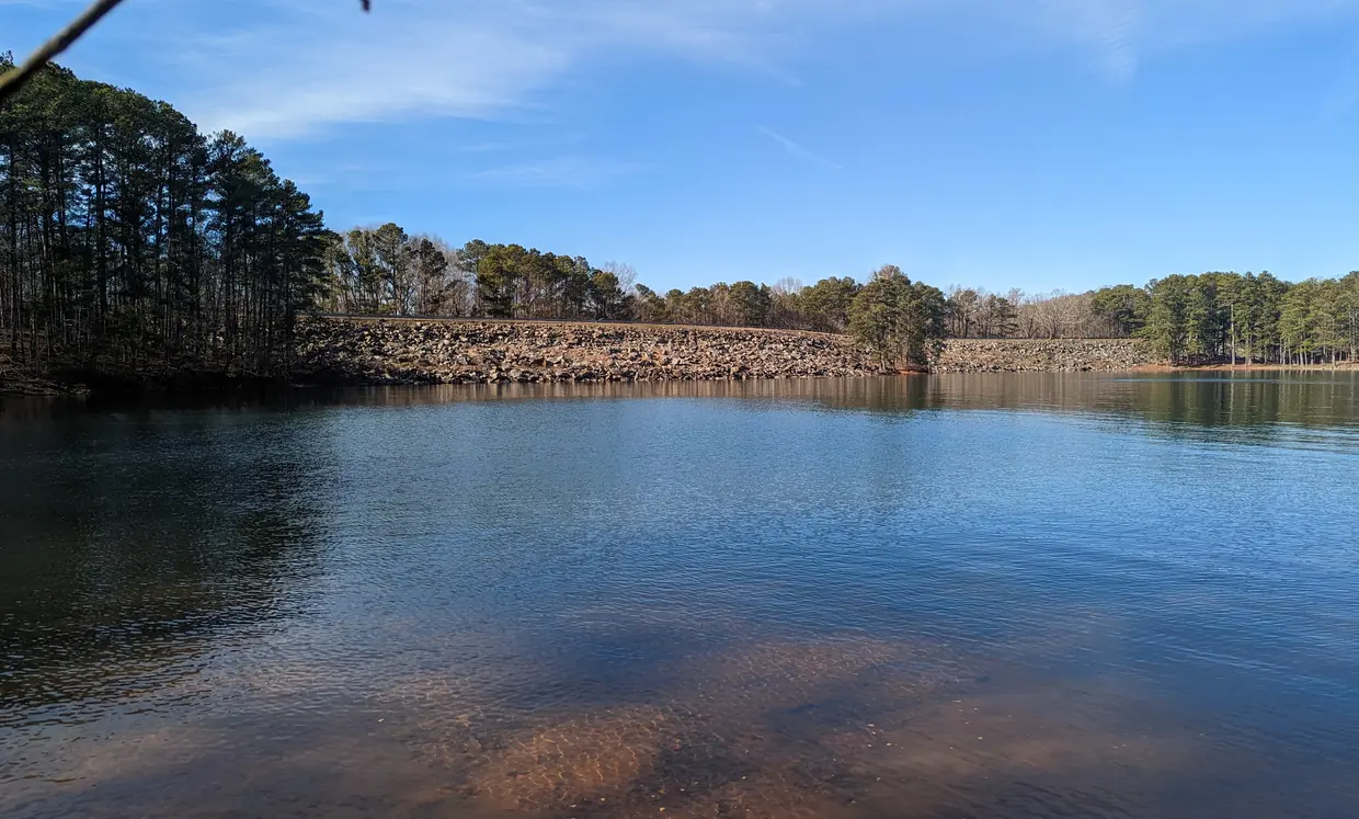 Clear rippled blue water over a sandy bottom, stretching to a long embankment faced with tan riprap boulders and topped by a road and pines.