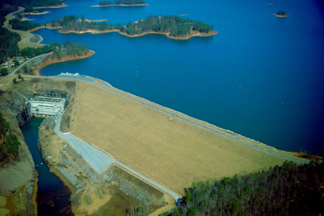 An aerial view of a long, straight earth dam with a grassy face holding back a deep blue lake scattered with wooded islands, a concrete powerhouse at its base.