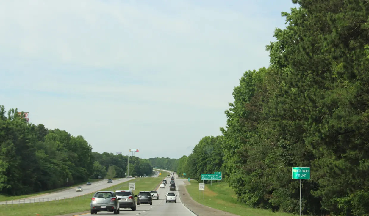 A green Flowery Branch city limit sign on the shoulder of a four-lane divided interstate lined with tall pines, cars heading toward an exit sign for Spout Springs Road.