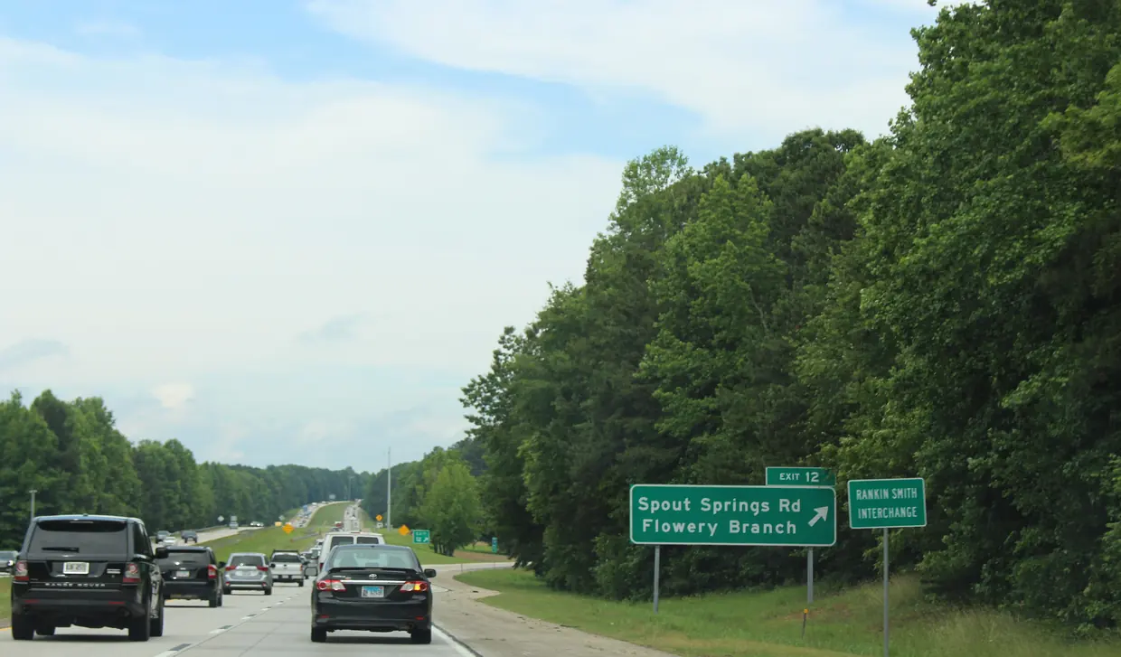 Cars on a multi-lane interstate passing a green sign for Exit 12, Spout Springs Road, Flowery Branch, against a wall of green trees.