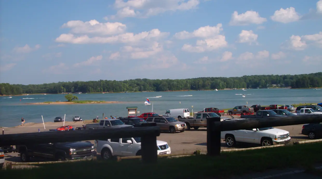 Pickup trucks with boat trailers parked above a boat ramp that slopes into a broad blue lake dotted with motorboats and a small sailboat.