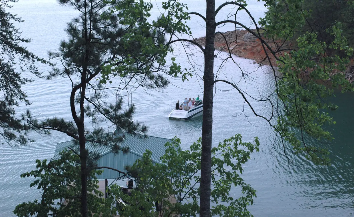 A white motorboat crossing pale blue-green water past a green metal boathouse roof, seen from a wooded hillside, with a red-clay point on the far shore.