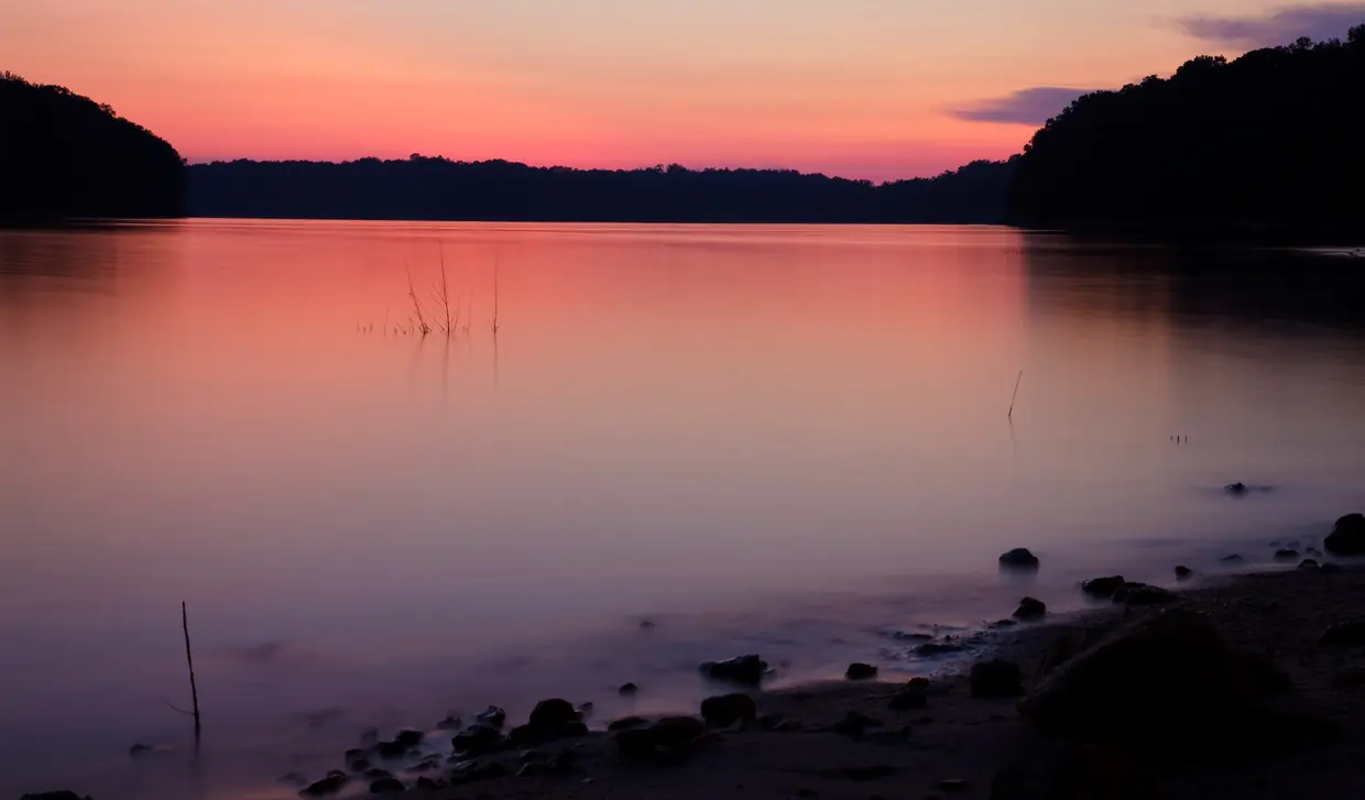 Glassy pink and violet water in a cove at dusk under an orange-to-pink sky, dark wooded hills on both sides and smooth dark stones on a sandy beach in front.