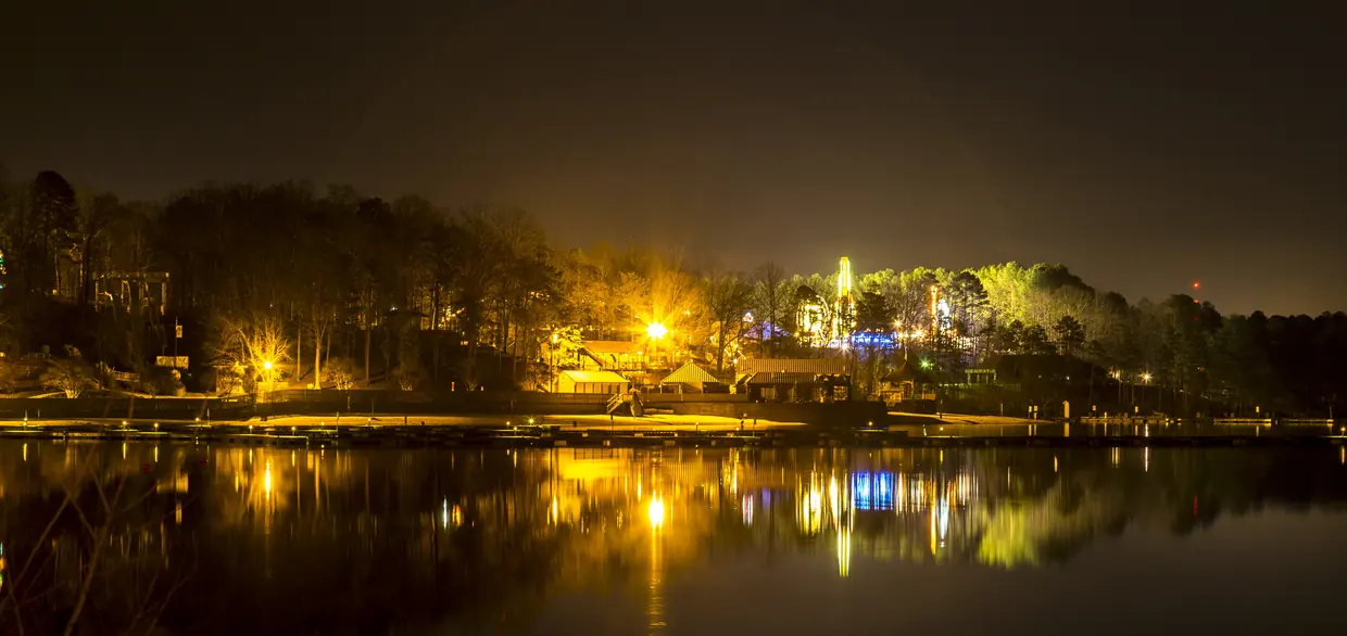 A wooded shoreline lit by warm orange lamps and low buildings at night, every light mirrored in perfectly still water.