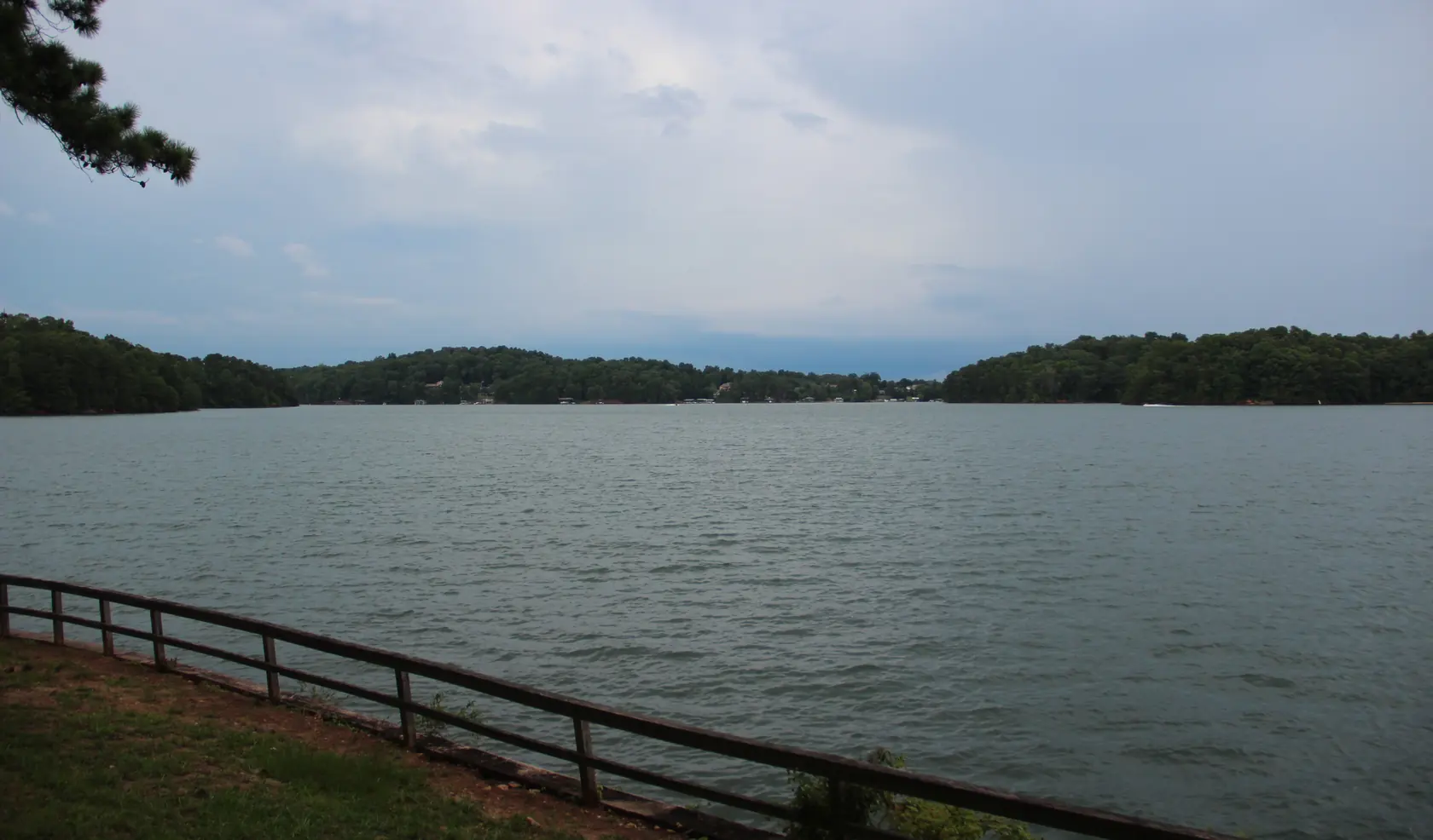 Wind-ruffled grey-green lake water stretching to low wooded hills dotted with houses and boat docks, under a pale overcast sky, with a wooden rail fence on a grassy bank in front.