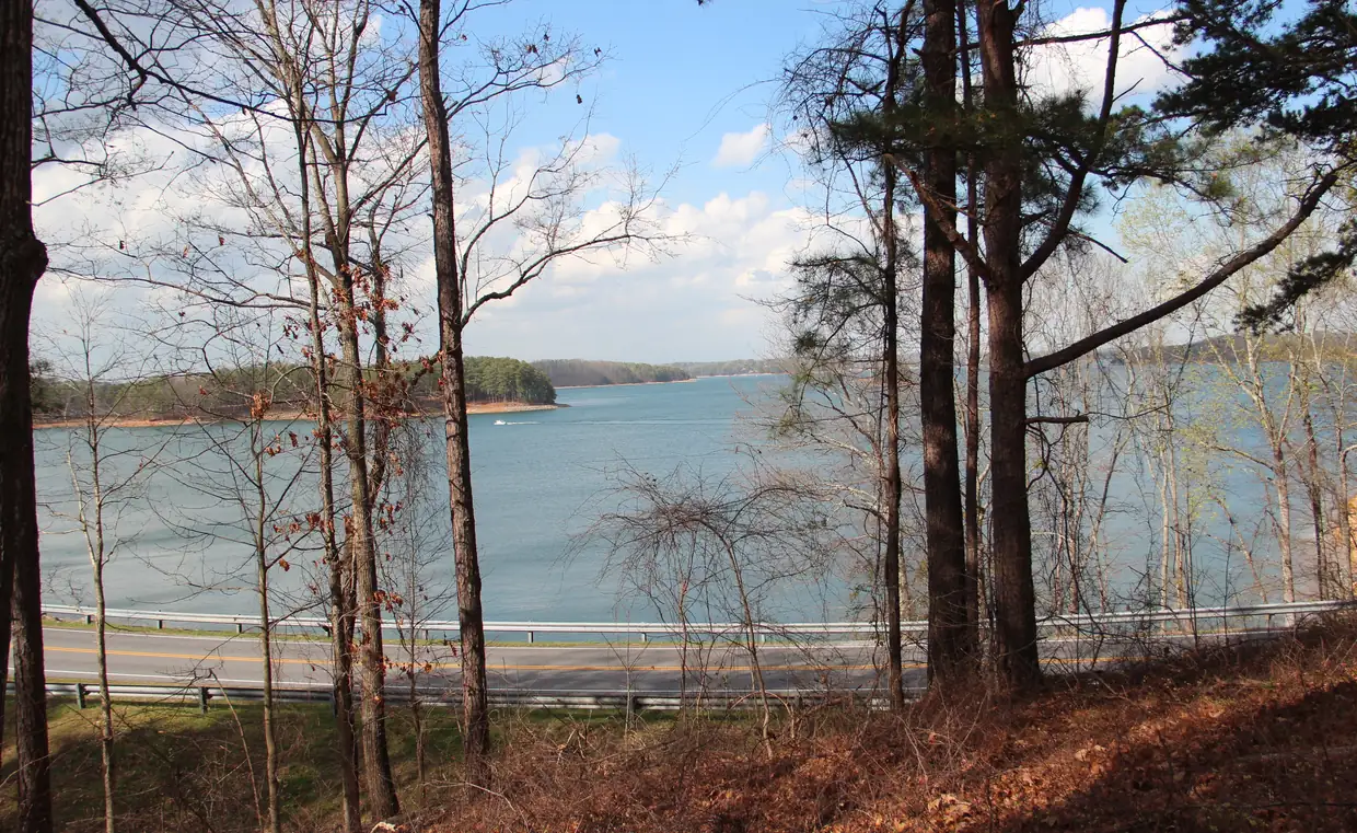 A wide blue arm of the lake seen through bare early-spring trees, opening toward wooded points with red-clay shorelines, one boat leaving a white wake.
