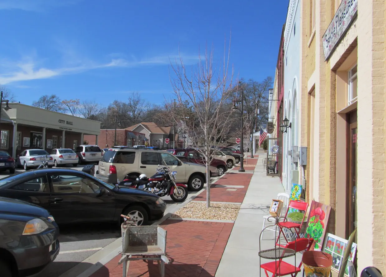 A sunny brick sidewalk past cream and pale-blue brick storefronts with arched windows and an American flag, angle-parked cars, and a low brick building signed City Hall across the street.