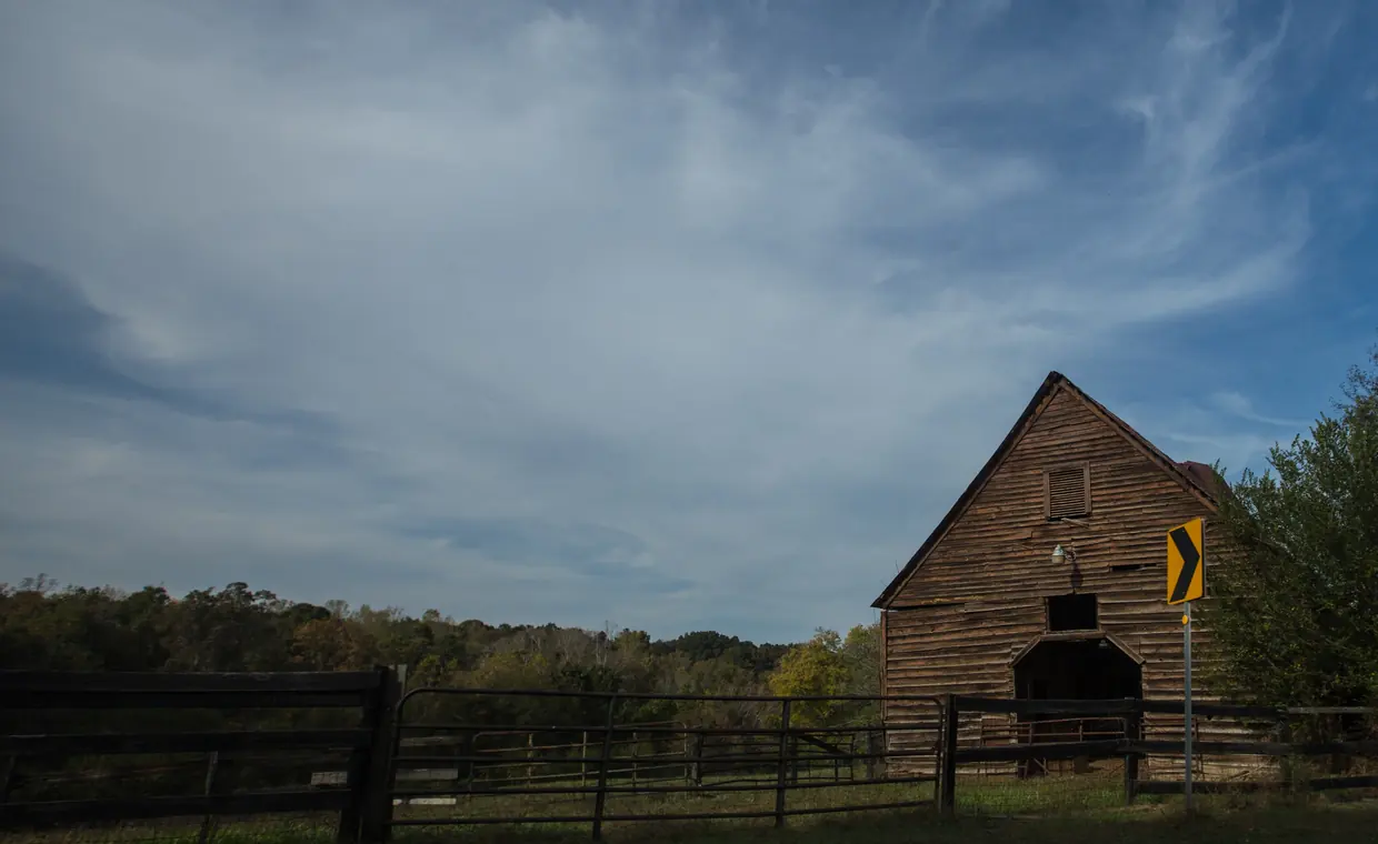 A weathered wooden barn at the edge of a pasture behind black board fencing, a wooded ridge with early autumn colour along the horizon under a wide sky.