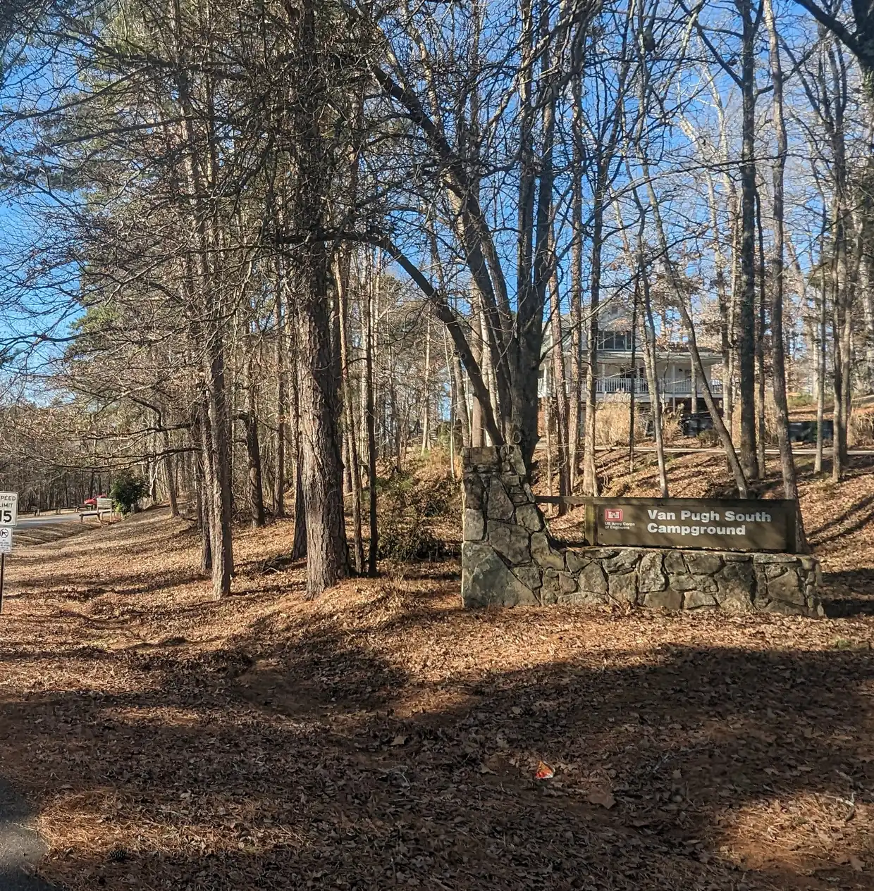 A stone-pillared wooden sign for Van Pugh South Campground with a U.S. Army Corps of Engineers emblem, beside a curving park road among tall pines.